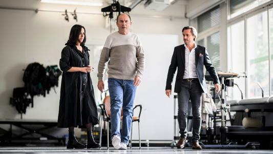 Three people, including a man and a woman, standing in a room discussing the effects of Parkinson's disease and spinal implant advancements.